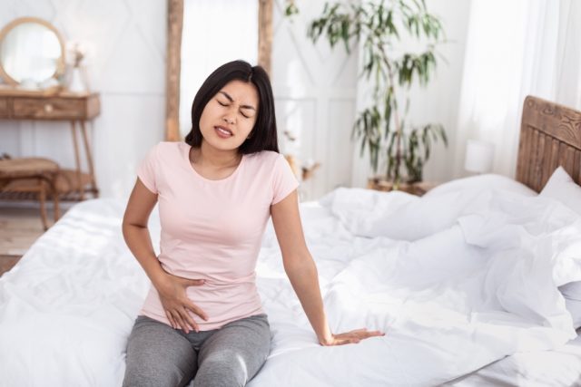 Woman sitting on her bed experiencing abdominal pain.
